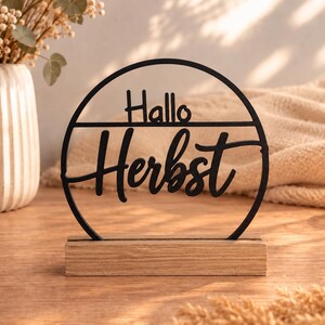 May include: A black circular decorative sign featuring the text "Hallo Herbst" in a mix of block and script lettering. The sign is mounted on a rectangular light-coloured wooden base, creating a stable stand for this tabletop display piece.