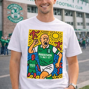 May include: White t-shirt featuring a colorful graphic of a soccer player in a green and white jersey with the text "Marathon Bet". The shirt is worn by a person in front of a stadium.