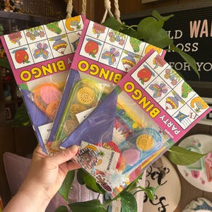 Könnte beinhalten: Drei Bingo-Party-Geschenkpakete mit farbenfrohen Illustrationen von Früchten, Schmetterlingen und Dinosauriern. Jedes Paket enthält eine Bingo-Karte und verschiedene Partygeschenke. Die Pakete werden in einer Hand gehalten.