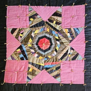 May include: A handmade quilt featuring a central star pattern constructed from multi-colored fabric strips in shades of blue, yellow, brown, and black. The star is surrounded by solid pink squares and black fabric panels, all secured with visible yellow yarn ties.