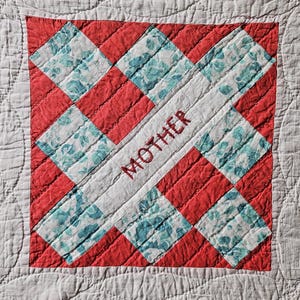 May include: Close-up of a handmade quilt with a central square design. The square features red and floral patterned fabric, with the word "MOTHER" embroidered in red. The quilt has a white border and a textured, light orange background.