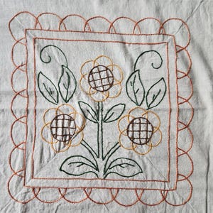 以下が含まれることがあります： 明るい色の生地に刺繍された花の模様。茶色と黄色の中心、緑の葉と茎を持つ3つのヒマワリが特徴です。正方形のデザインは、オレンジ色の糸でスカラップのボーダーで縁取られています。
