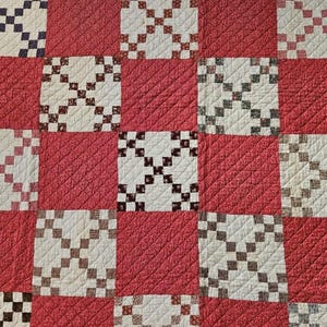May include: A close-up of a red and white quilt with a checkerboard pattern. The quilt features alternating squares of red fabric and white squares with a cross-stitch design in various colours, including black, brown, and pink.