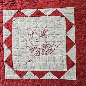 Könnte beinhalten: Ein rot-weißer Quilt mit einem quadratischen Block, der in der Mitte einen gestickten Vogel auf einem Ast zeigt. Das Quadrat ist von einer rot-weißen Sägezahnkante umrahmt, und die gesamte Stoffoberfläche weist ein gestepptes Gittermuster auf.