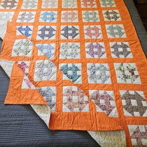 May include: A patchwork quilt featuring a grid of orange fabric sashing separating individual square blocks. Each block contains a multi-colored patterned fabric in a geometric design. The quilt is folded at one corner, revealing a light-colored backing.