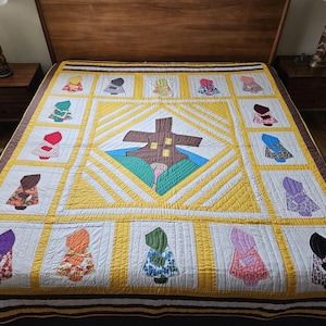 Op de afbeelding: Een handgemaakte quilt met een centraal geometrisch ontwerp en zich herhalende figuren in verschillende kleuren. De quilt heeft een bruin, geel en wit kleurenschema, met een rand van bruine en gele strepen. De figuren zijn gekleed in kleurrijke outfits.
