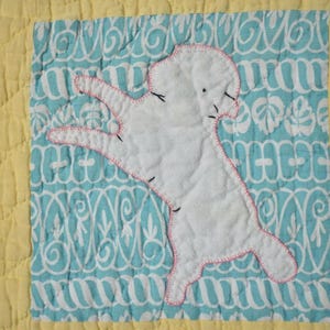Puede incluir: Un edredón hecho a mano con una aplicación blanca de un perro con costuras rosas sobre un fondo estampado en azul y blanco. El edredón tiene un borde amarillo con un diseño acolchado.