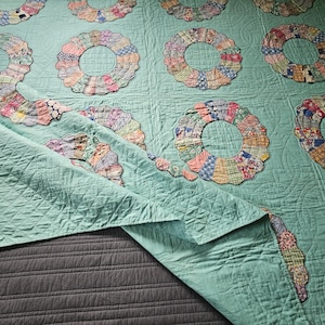 Puede incluir: Una colcha de color verde menta claro con un patrón repetitivo de parches florales circulares de bordes festoneados en varios colores. La tela tiene una textura acolchada con líneas de costura visibles y una esquina está doblada.