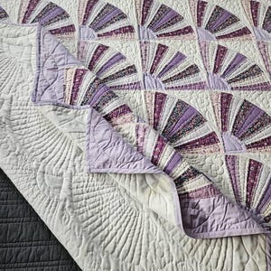 Peut inclure: Une courtepointe faite à la main avec un motif en éventail dans les tons de lavande, de violet et de blanc. La courtepointe présente un motif cousu détaillé sur le fond blanc et une bordure lavande. La courtepointe est pliée, révélant les coutures complexes.