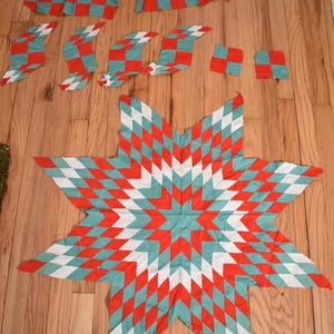 May include: A large, eight-pointed star quilt block featuring a geometric pattern of diamond-shaped fabric pieces in red, teal, and white. Several smaller, detached strips and squares of the same red, teal, and white fabric are arranged above.
