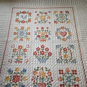 May include: A white quilt featuring a grid of twelve embroidered squares with colourful floral, bird, and heart motifs in red, yellow, blue, and green. The fabric has a quilted texture with a scalloped red border surrounding the central design.