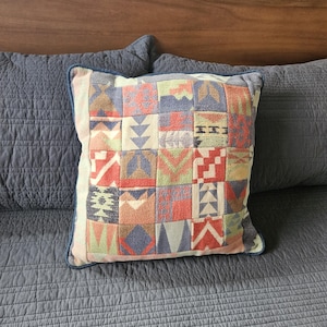 Könnte beinhalten: Quadratisches Zierkissen mit einem Patchwork-Design in Rot-, Blau-, Grün- und Beigetönen. Das Kissen hat einen blauen Rand und liegt auf einer grau gesteppten Tagesdecke. Das Design des Kissens zeigt geometrische Formen.
