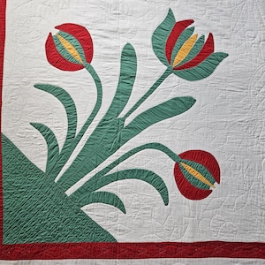 Op de afbeelding: Een handgemaakte quilt met een bloemenontwerp. De quilt toont rode, groene en gele tulpachtige bloemen tegen een witte achtergrond. De randen zijn afgezet met een rode streep. De quilt heeft een gewatteerde textuur.