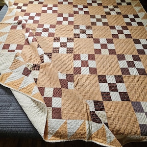 Può includere: Un copriletto trapuntato con un design patchwork di quadrati alternati color marrone chiaro, bianco e marrone scuro. La trapunta presenta un motivo a griglia ripetuto con cuciture visibili e un angolo è ripiegato per rivelare il lato inferiore color crema.