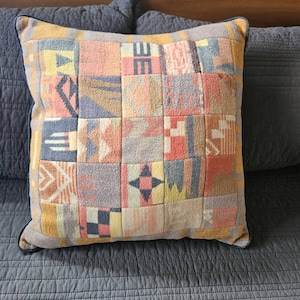 Puede incluir: Cojín decorativo cuadrado con diseño de patchwork. El cojín presenta una variedad de patrones geométricos en tonos amarillos, rosas, azules y grises. Tiene un borde azul oscuro y está sobre una colcha azul.