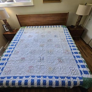 Könnte beinhalten: Eine weiße Steppdecke mit gestickten Blumen- und Tiermotiven in der Mitte, umgeben von einem blau-weiß karierten Rand mit grünem Besatz. Die Decke liegt auf einem Bett mit hölzernem Kopfteil, das von zwei Nachttischlampen flankiert wird.