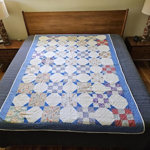 May include: A handmade quilt with a blue border and white backing is displayed on a bed. The quilt features a repeating pattern of white circles, blue squares, and floral print patches. The bed has a wooden headboard and two matching bedside tables.