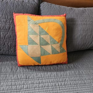 Op de afbeelding: Vierkantig decoratief kussen met een levendige oranje achtergrond en een patchwork ontwerp met driehoeken en een gebogen vorm in groen- en beigetinten. Het kussen heeft een rode rand.