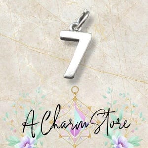 Könnte beinhalten: Ein silberfarbener Charm-Anhänger mit der Zahl 7 und einer Schlaufe zum Befestigen an einem Armband oder einer Halskette. Der Anhänger ist vor einem marmorierten Hintergrund mit dem Schriftzug "A Charm Store" platziert.