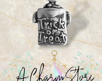 May include: A silver-toned charm in the shape of a t-shirt, with the words "Trick or Treat" engraved on the front. The charm has a loop at the top for attaching to a bracelet or necklace. The background is a light, patterned design.