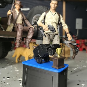Könnte beinhalten: Ein schwarzes Spielzeug-Panzerfahrzeug steht auf einer blauen Box neben einer goldenen Trophäe. Im Hintergrund befinden sich zwei Actionfiguren, eine mit einem Gewehr und die andere mit einer Fernbedienung. Ein Golden Retriever ist ebenfalls vorhanden.