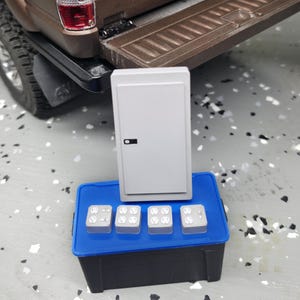 Puede incluir: Un panel eléctrico gris en miniatura con un asa negra y cuatro enchufes en un contenedor azul. El panel está sobre un contenedor negro y azul, con una caja de camión marrón en el fondo.