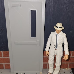 May include: A gray door with a rectangular window and a silver handle stands next to a figure in a white suit, fedora, and tie. The figure is standing on a surface of small white and gray pieces, with a brick wall in the background.
