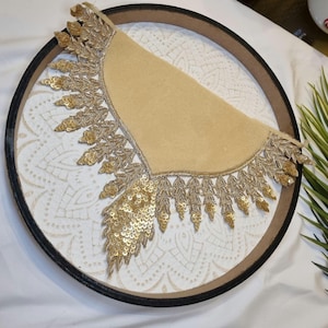 Op de afbeelding: Een ronde, decoratieve dienblad met een crèmekleurig, patroon oppervlak en een zwarte en bruine rand. Een beige, halfrond stoffen stuk is versierd met gouden pailletten en bladvormige versieringen.