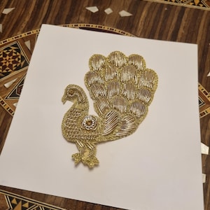 May include: A gold and silver embroidered peacock applique on a white background. The peacock has a detailed, textured design with a fan-shaped tail. A small, round jewel is on the peacock's body. The applique is likely for clothing or craft projects.