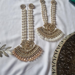 May include: Two ornate, gold-toned earrings with pearl strands and a fan-shaped base. The earrings have a floral design at the top and a fringe of peach-colored beads at the bottom. The earrings are displayed on a white surface.