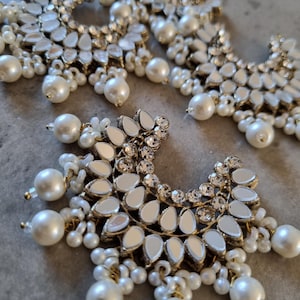 May include: Close-up of several ornate, silver-toned earrings. Each earring features a crescent shape with mirrored accents, surrounded by small, white pearls and larger, dangling pearls. The earrings have a delicate, elegant design.