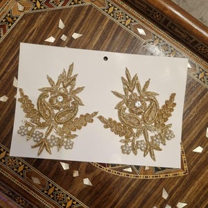 May include: Two gold embroidered floral appliques with clear rhinestone centers and small pearl accents. The appliques are on a white card, set against a decorative wooden tray.