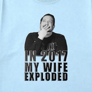 May include: Light blue t-shirt with a black and white image of a man and the text "IN 2017 MY WIFE EXPLODED". The shirt is made of a soft material and has a crew neck.