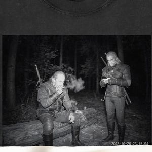 May include: Black t-shirt featuring a black and white image of two men in a forest setting. One man is smoking, while the other is looking at a device. The shirt has a tag that says LUMEDSHIRE.