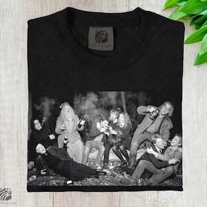 May include: Black t-shirt with a black and white image of a group of people in a forest. The image depicts people drinking, smoking, and socialising. The shirt has a Lumenshire logo.