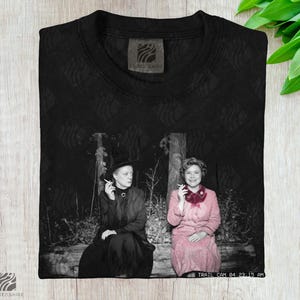 May include: Black t-shirt featuring a black and white image of two women smoking. One woman wears a black dress and hat, the other a pink dress. The shirt has the text "TRAIL CAM 04:23:15 AM" and the brand name "LUMENSHIRE".