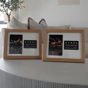 May include: Two framed photos with light wood frames sit on a gray table. Each frame contains a photo and a black and white label that reads "HYROX MANCHESTER". The background includes a white wall, a couch, and a decorative plant.