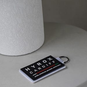 May include: A black and white HYROX Cardiff keyring with a metal ring. The text "HYROX CARDIFF" is displayed vertically. The bottom of the keyring reads "OPEN_DOUBLES_SEASON_2025/2026".