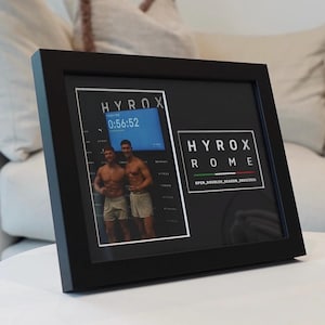 May include: Black framed photo featuring two men and the text "HYROX ROME". The photo includes a digital display showing a time of 0:56:52. The frame is on a white surface.