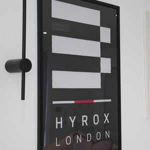 May include: Black framed poster with white and black geometric design, and the text "HYROX LONDON". A black, minimalist wall-mounted light fixture is to the left of the poster.