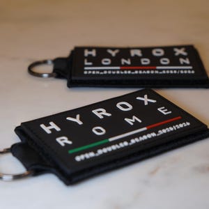 May include: Two black rectangular keychains with white text. One says "HYROX LONDON" with a red stripe, the other says "HYROX ROME" with a green and red stripe. Both have a metal keyring.