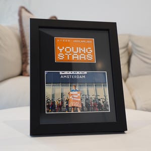 May include: A framed photograph featuring the text "YOUNG STARS" in orange pixelated letters. Below, a photo shows a person holding a sign with the same text in front of bicycles. The frame is black and sits on a white surface.