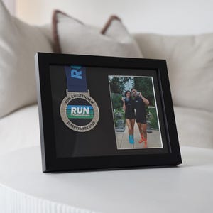 May include: Black framed display featuring a running medal and a photograph. The medal is silver with a blue ribbon and the text "RUN Cheltenham 21 September 2025". The photo shows two people in running attire.