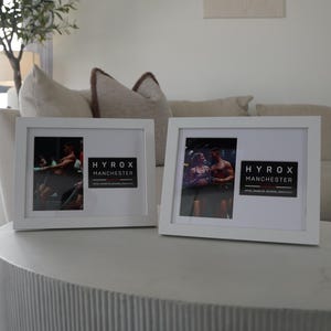 Op de afbeelding: Twee witte fotolijsten met foto's en de tekst "HYROX MANCHESTER". Ze staan op een witte, getextureerde tafel. De achtergrond bevat een bank en decoratieve elementen.