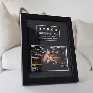 May include: Black framed print with the text "HYROX BIRMINGHAM" and a race image. The print includes the names Gregor Johnston and Hattie Campbell, and the time 1:11:0. The frame is displayed on a white surface.