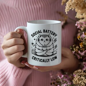 May include: White ceramic mug featuring a black and white cat illustration wrapped in a blanket, holding a cup. The text on the mug reads "SOCIAL BATTERY CRITICALLY LOW". The mug has a white handle and is held by a person.