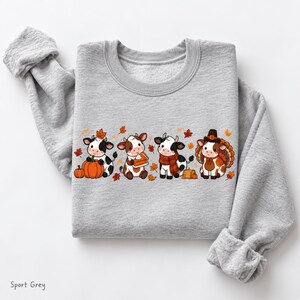 May include: A light grey crewneck sweatshirt featuring a graphic print of four cartoon cows. One cow sits with a pumpkin, another holds a slice of pie, one wears a scarf, and the fourth is dressed as a turkey. The design includes scattered autumn leaves.