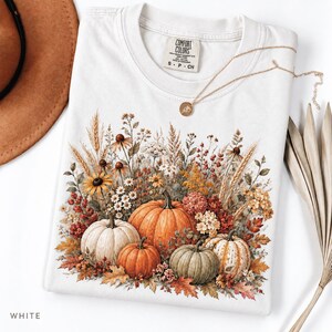 May include: A white t-shirt featuring a graphic print of various pumpkins and autumn wildflowers including wheat, daisies, and berries. The shirt has a Comfort Colors brand label at the neck and is accompanied by a thin gold-toned chain necklace.