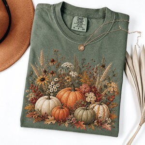 May include: An olive green t-shirt featuring a graphic print of four pumpkins surrounded by wheat, wildflowers, and autumn leaves. The shirt has a Comfort Colors brand label at the neckline and is displayed with a gold necklace chain.