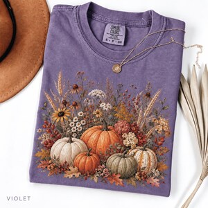 May include: A violet short-sleeved t-shirt featuring a graphic print of four pumpkins surrounded by autumn flowers, wheat stalks, and dried leaves. The shirt has a crew neckline and a label printed on the inside neck area with the text Comfort Colors.